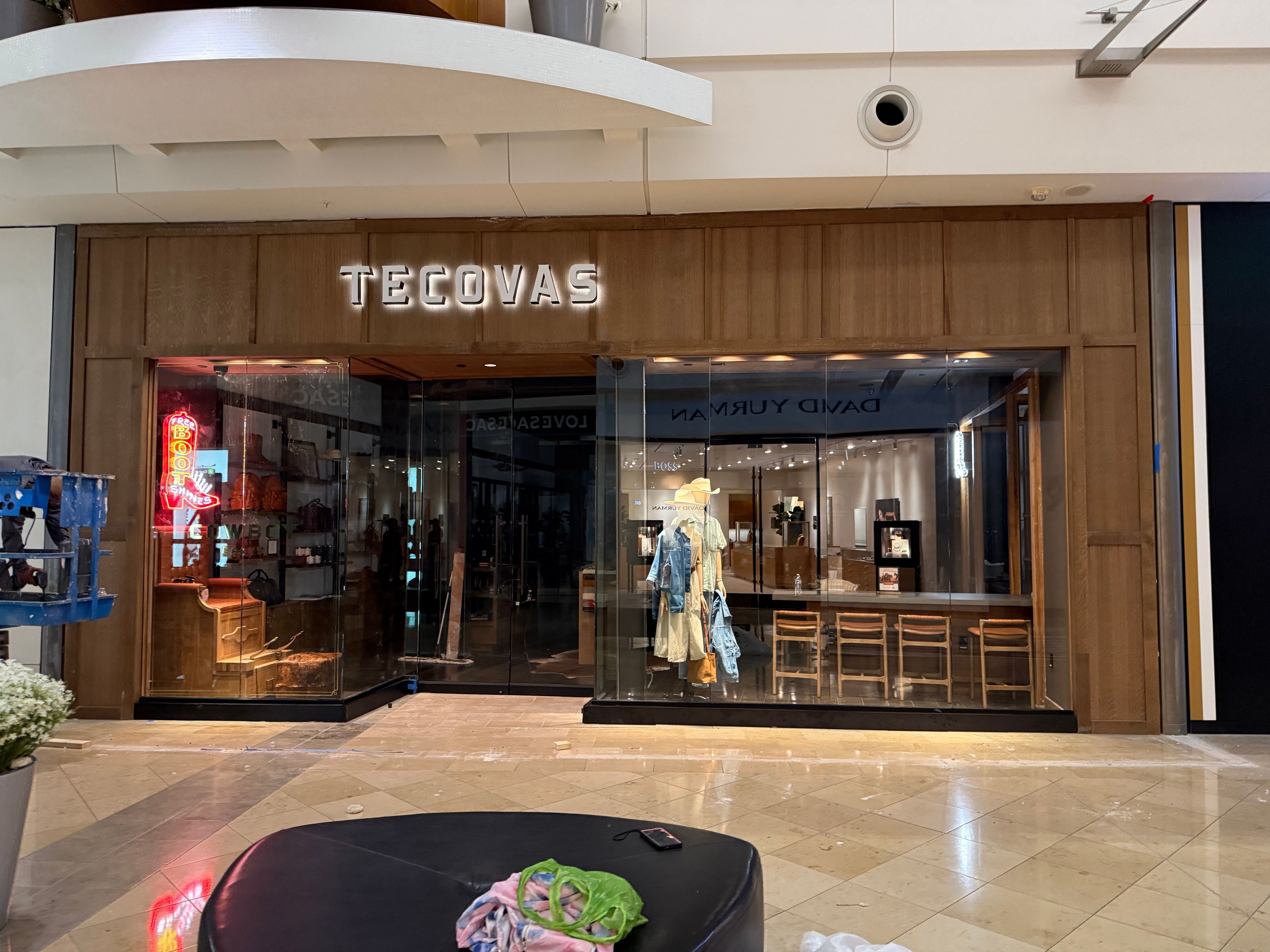 A Tecovas retail store with glass windows displays boots, hats, and clothing; the interior includes a seating area and wooden decor.