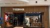 A Tecovas retail store with glass windows displays boots, hats, and clothing; the interior includes a seating area and wooden decor.