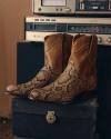 A pair of brown and snakeskin cowboy boots sit on a black box, with a vintage stereo system in the background.