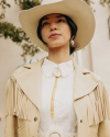 Person wearing a large cowboy hat, fringed jacket, and bolo tie, standing outdoors.