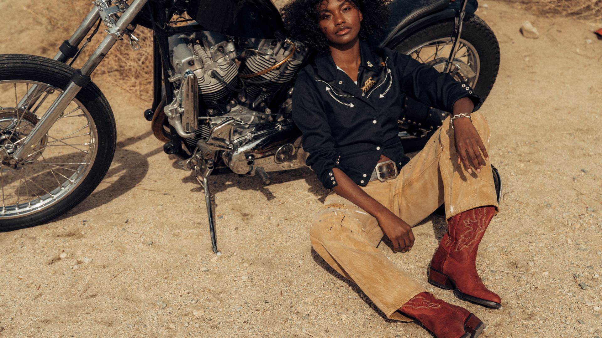 A person wearing a black shirt, tan pants, and red boots sits on the ground next to a motorcycle in a desert setting.