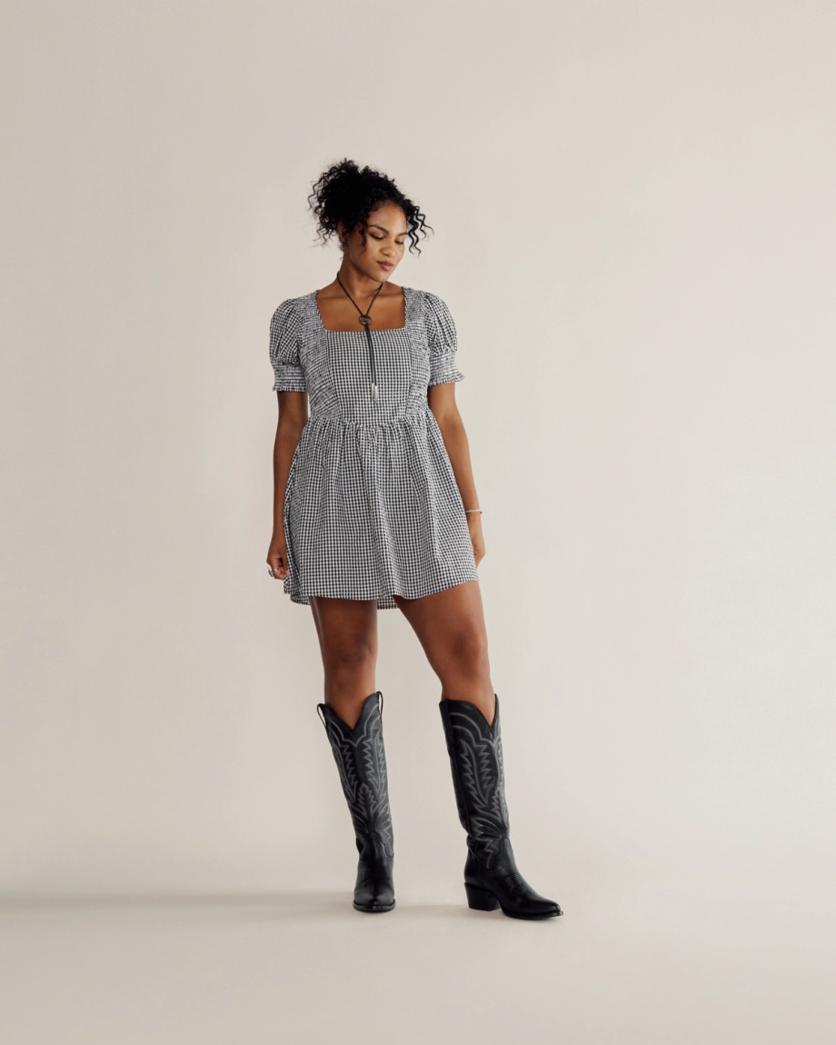 A woman in a black and white checkered dress and black cowboy boots stands against a plain light background, looking slightly downward to her left.