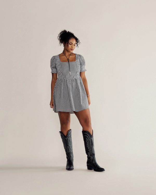 A woman in a black and white checkered dress and black cowboy boots stands against a plain light background, looking slightly downward to her left.