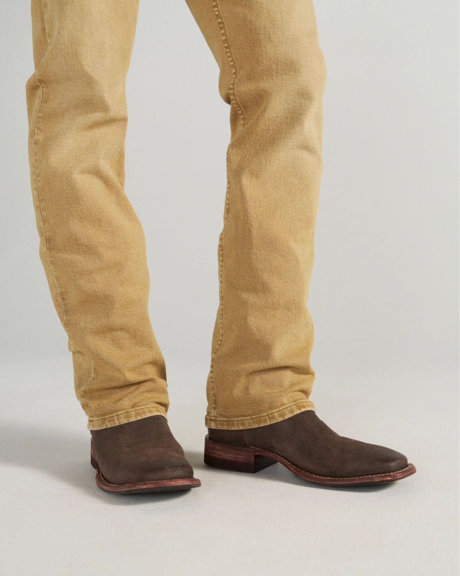The Doc in Moss Suede. A person wearing tan jeans and dark brown leather cowboy boots stands on a light-colored floor, their style elevated by the subtle texture of Moss Suede.