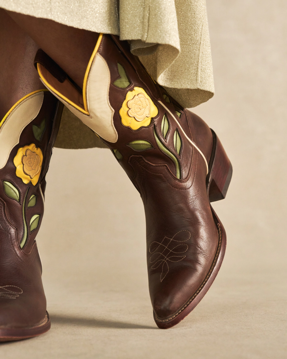 The Jolene in Truffle Cowhide. Close-up of a person wearing brown truffle cowhide cowboy boots with yellow floral and green leaf designs, standing on a neutral background.