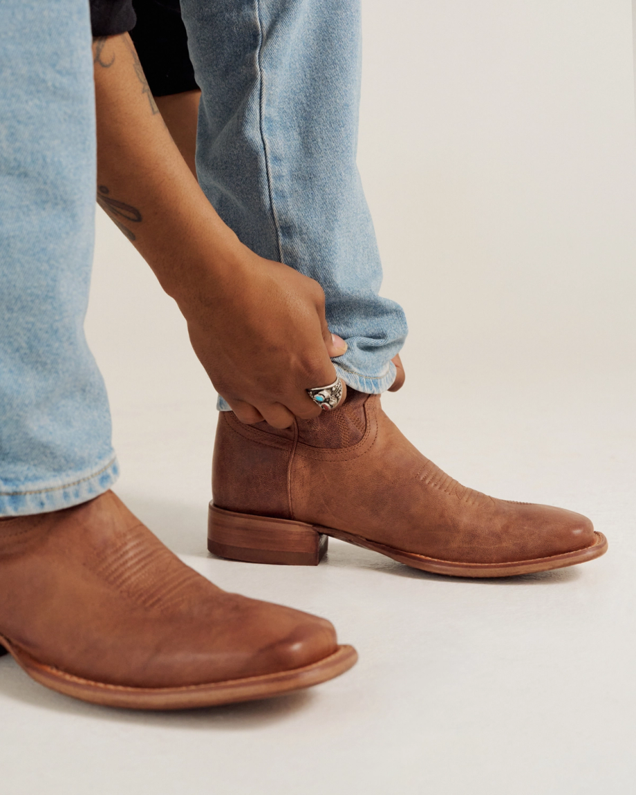 A person wearing light blue jeans adjusts the cuff of their pant leg above a brown leather ankle boot.