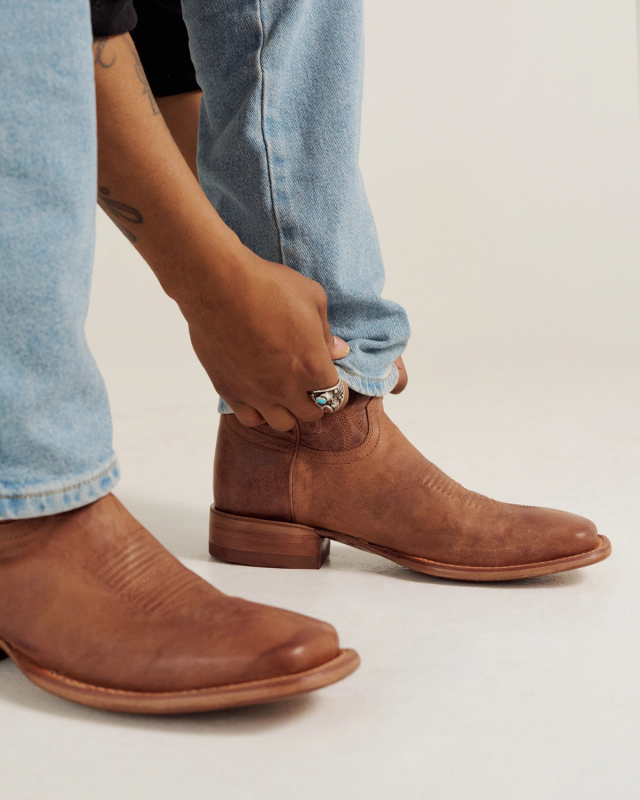 A person wearing light blue jeans adjusts the cuff of their pant leg above a brown leather ankle boot.