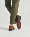 The Bennett in Saddle Alligator. A person wearing cuffed olive green pants and brown Saddle Alligator leather loafers is stepping forward, showing the sole of one shoe against a plain background.