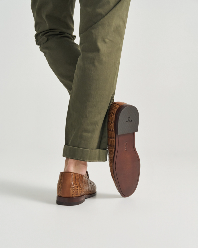 The Bennett in Saddle Alligator. A person wearing cuffed olive green pants and brown Saddle Alligator leather loafers is stepping forward, showing the sole of one shoe against a plain background.