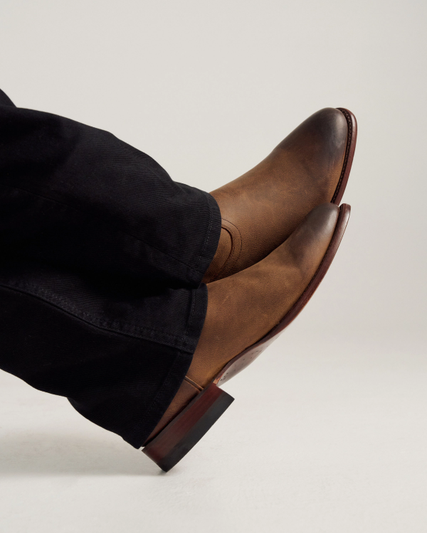 A person wearing black pants and brown leather dress boots with a slight heel, shown from the side against a plain background.