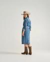 A woman wearing a denim dress, cowboy hat, and brown boots stands facing backward, looking over her shoulder against a plain background.