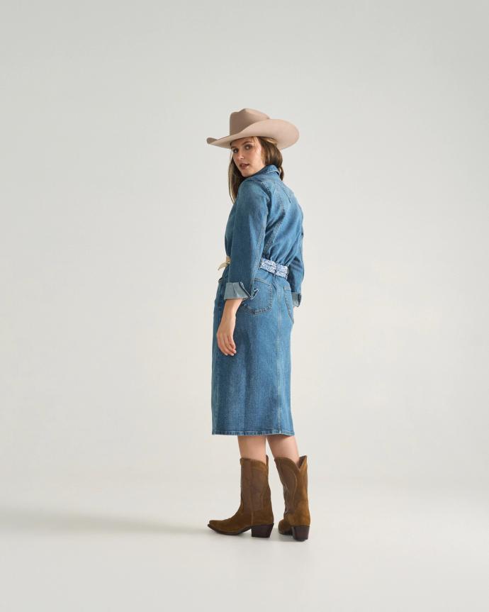 A woman wearing a denim dress, cowboy hat, and brown boots stands facing backward, looking over her shoulder against a plain background.