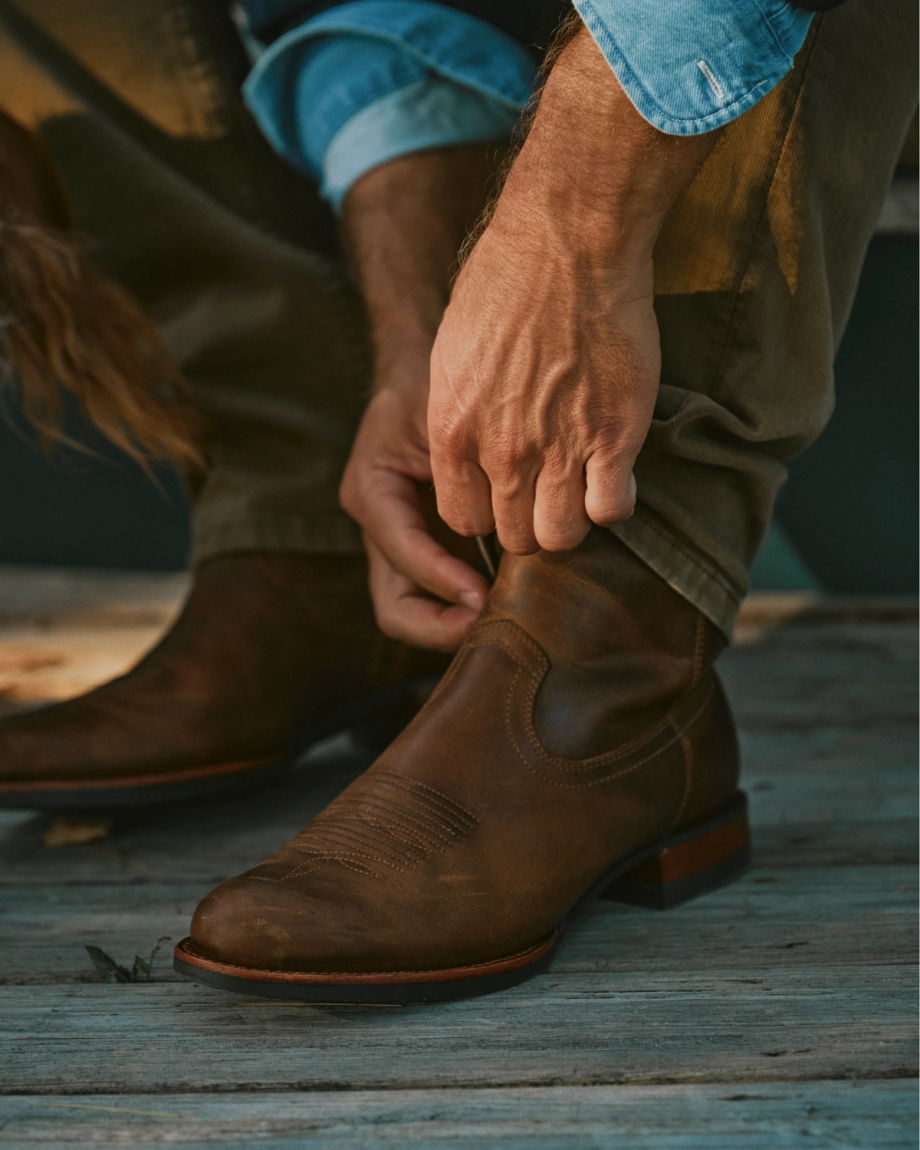 The Wayne in Maplewood Cowhide. A person in khaki pants and a denim shirt pulls on The Wayne Cowhide brown leather cowboy boot while sitting on a wooden surface.