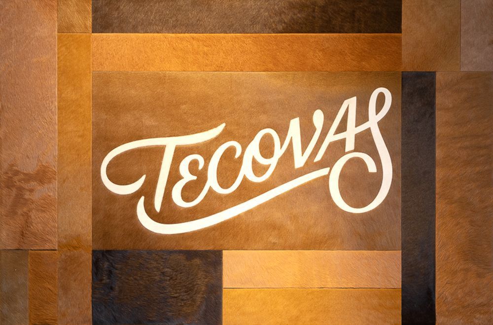 White script logo reading 'Tecovas' displayed on a background of rectangular brown and tan leather panels.