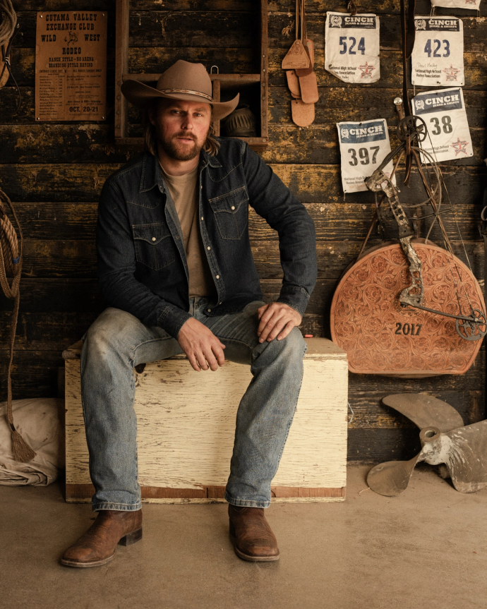 A man in a cowboy hat and denim jacket sits on a wooden box in a rustic setting with horse gear, award plaques, and a vintage fan nearby.