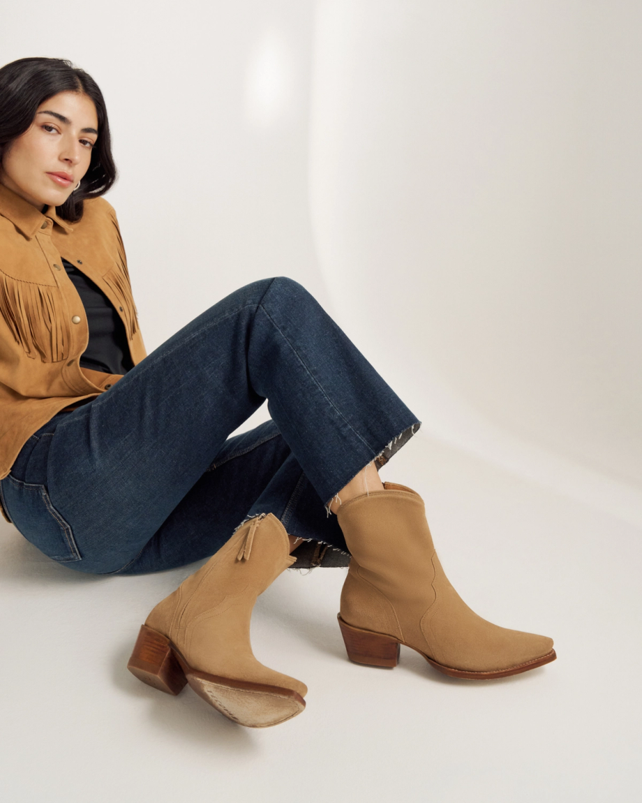 A woman in a tan fringe jacket, dark jeans, and tan suede boots sits on the floor against a plain light background.
