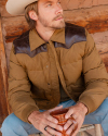 Man in a brown jacket and cowboy hat sits and holds a mug, looking to the side. Background features wooden elements.