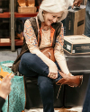 A woman sits on a bench, smiling as she tries on a brown ankle boot in a shoe store. A shopping bag rests on the floor beside her.