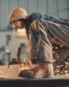 Person wearing protective clothing operates an angle grinder, producing sparks in a workshop setting. Focus is on the worker’s boots and flying sparks.