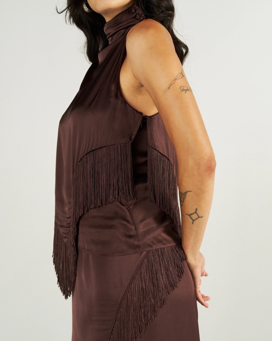 Woman wearing a sleeveless, brown satin dress with fringe details, standing sideways against a plain background. Visible tattoos on her arm.