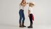 Two children in cowboy boots and jeans stand facing each other; one places a white cowboy hat on the other's head.