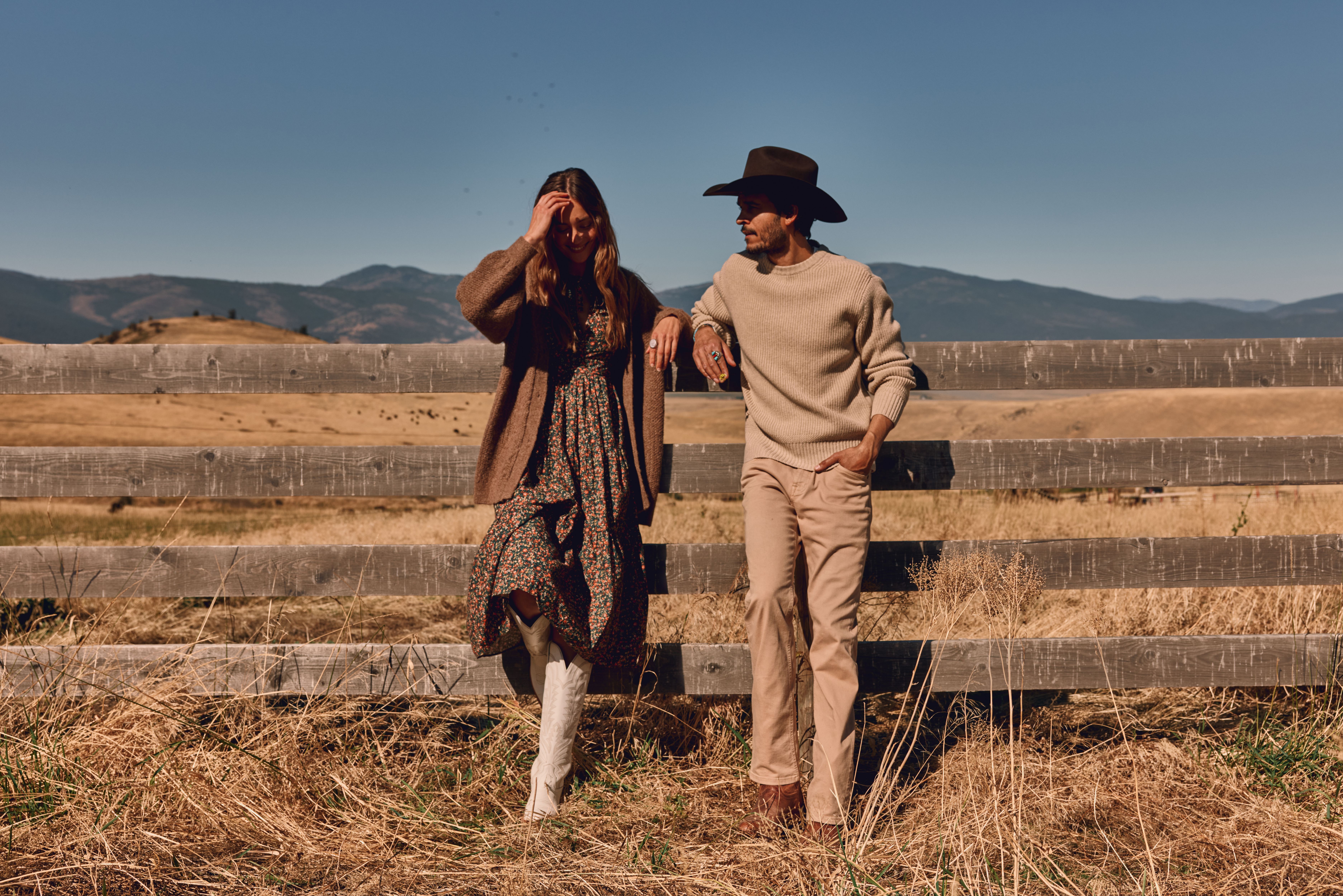 A woman in a floral dress and boots and a man in a hat and sweater stand by a wooden fence in a dry field with hills in the background.