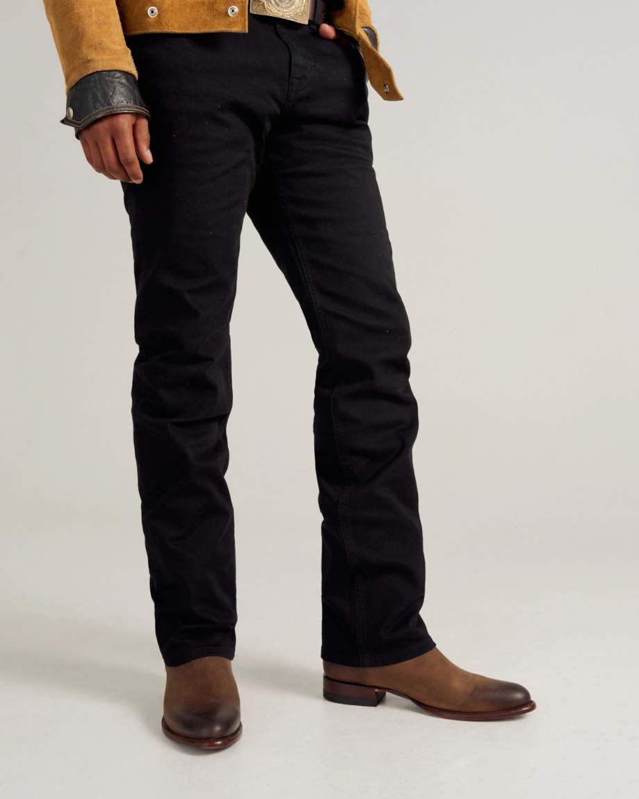 Person standing against a plain background wearing black jeans, brown leather boots, a mustard-colored jacket, and a black shirt sleeve partially visible.