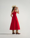 Woman in a red sleeveless dress, tan cowboy hat, and brown cowboy boots stands facing backward, looking over her shoulder against a plain background.