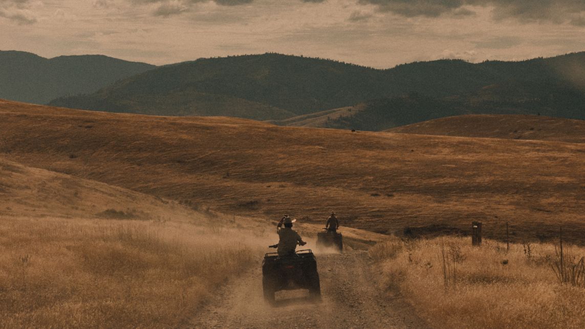 Two people ride ATVs on a dusty trail through rolling hills under a cloudy sky.