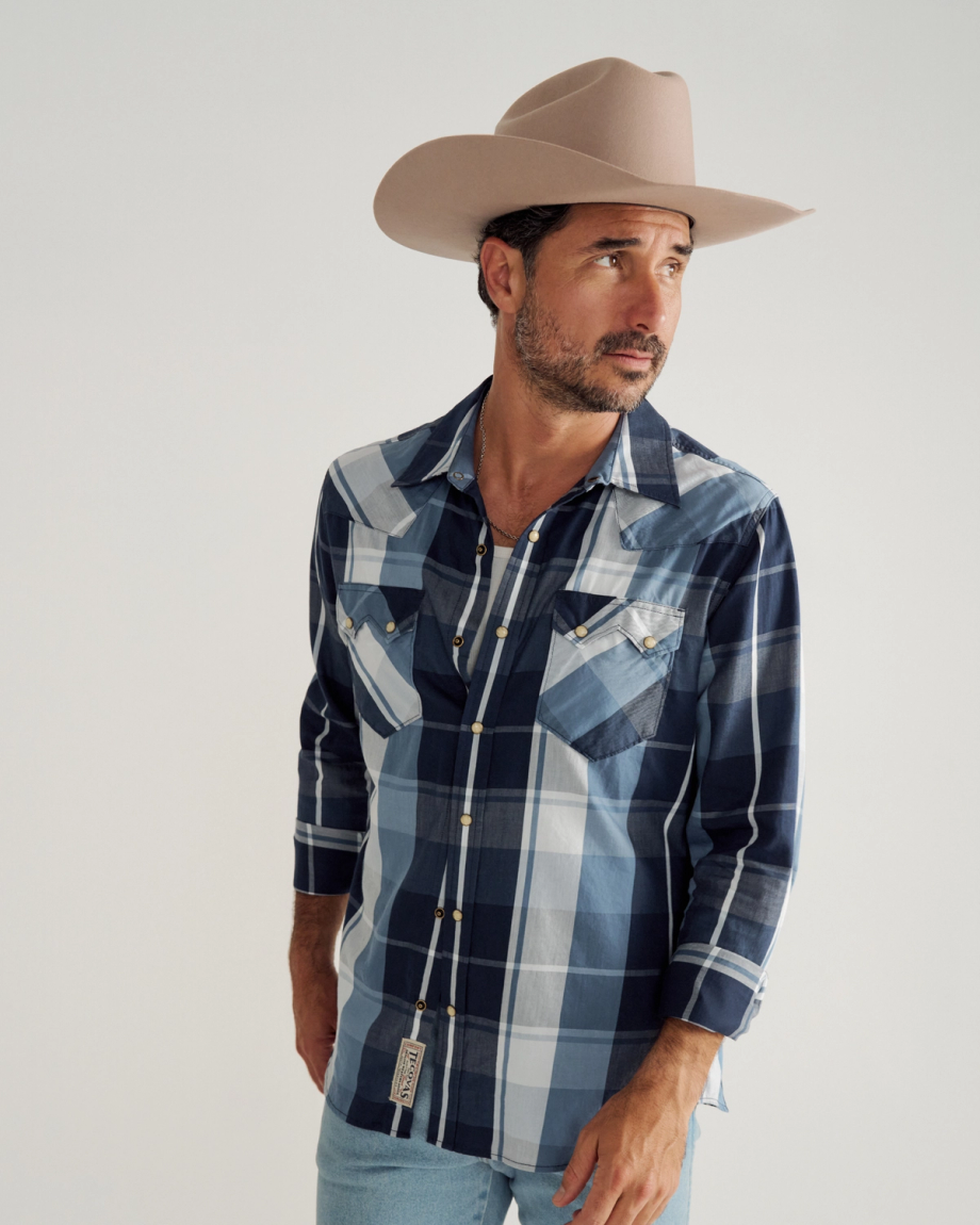 Man wearing a beige cowboy hat, blue plaid shirt, and light blue jeans stands against a plain white background, looking off to the side.