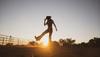 Silhouette of a person wearing a cowboy hat and boots, kicking up dust at sunset in a rural outdoor setting with fencing and trees in the background.