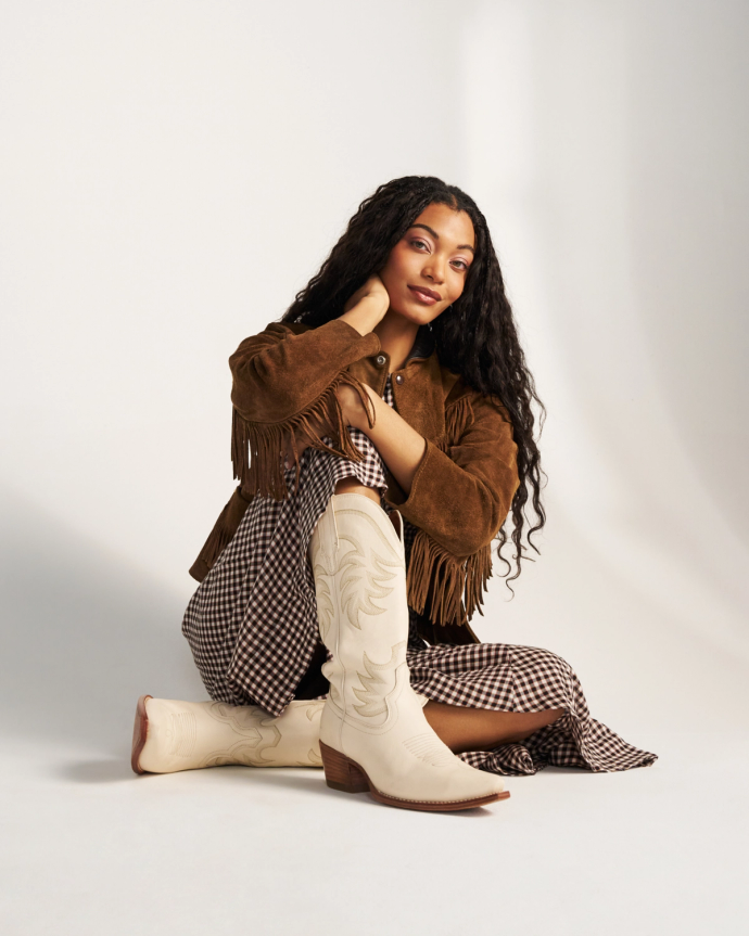 The Annie in Bone Cowhide. Woman sitting on the floor wearing a brown fringed jacket, checkered dress, and The Annie Cowhide white cowboy boots against a plain background.