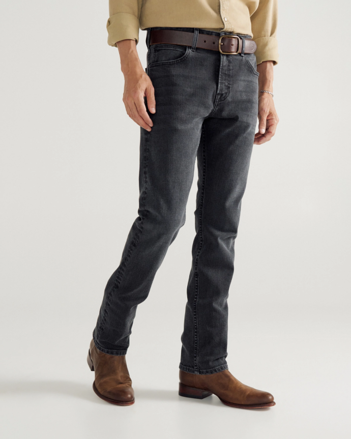 Person wearing a beige shirt, dark grey jeans with a brown belt, and brown leather boots, standing against a plain background.