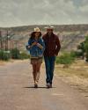 Two people wearing cowboy hats and boots walk side by side on a rural road with hills and cloudy skies in the background.