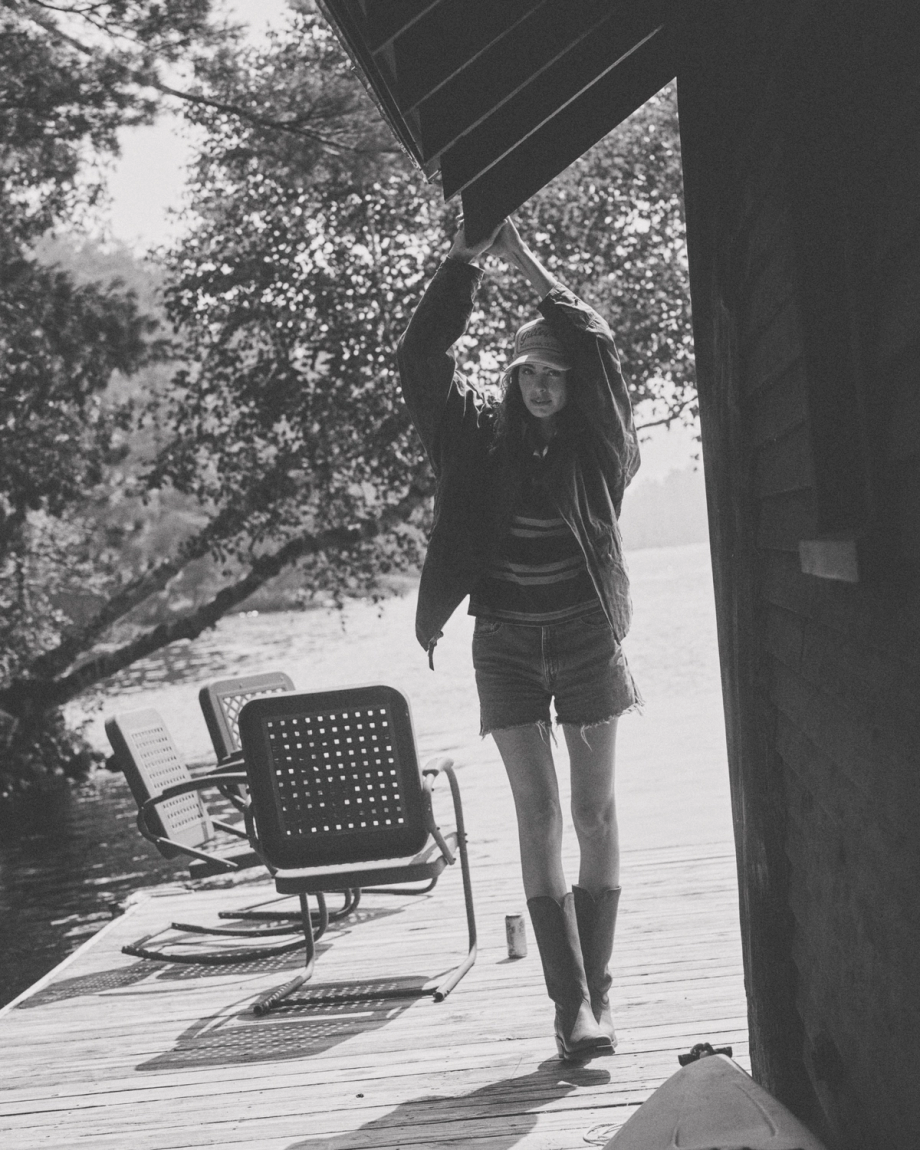 The Charlie in Scarlet Cowhide. A person in boots and a jacket stands on a lakeside dock, holding onto the roof edge. Scarlet Cowhide chairs and trees are visible in the background.