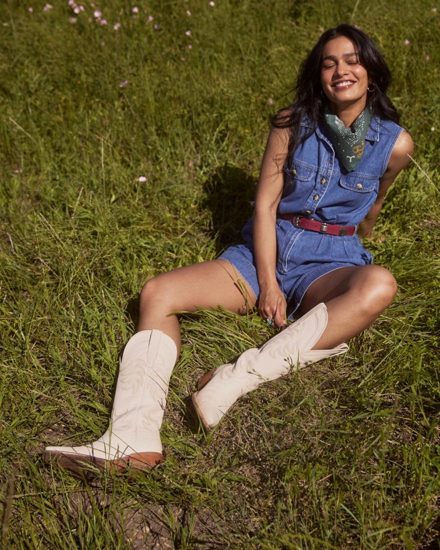Person sitting on grass wearing a denim outfit, white boots, and a green scarf, smiling with eyes closed.