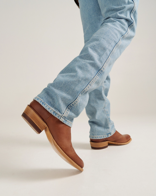 Person wearing light blue jeans and brown leather boots walking, with one foot lifted slightly off the ground against a plain background.