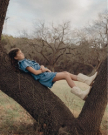 A young girl in a blue dress and white boots lies back on a tree branch in a grassy field, with bare trees in the background.