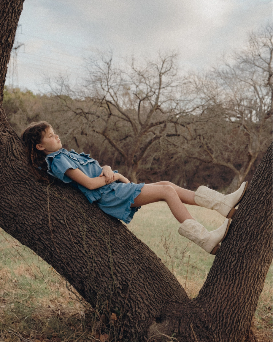 A young girl in a blue dress and white boots lies back on a tree branch in a grassy field, with bare trees in the background.