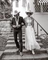A man in a striped suit and a woman in a long white dress, both wearing cowboy hats, walk arm in arm down outdoor steps beside a building.