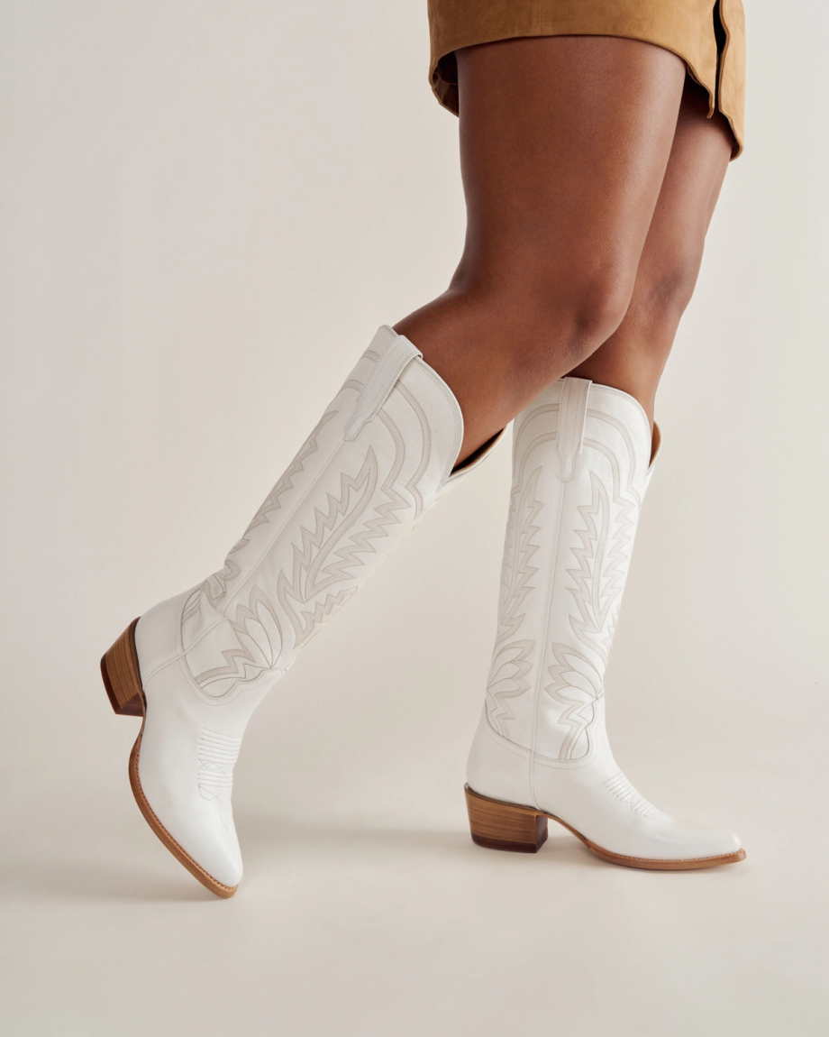 Person wearing tan fringed shorts and white embroidered knee-high cowboy boots, standing against a plain background.