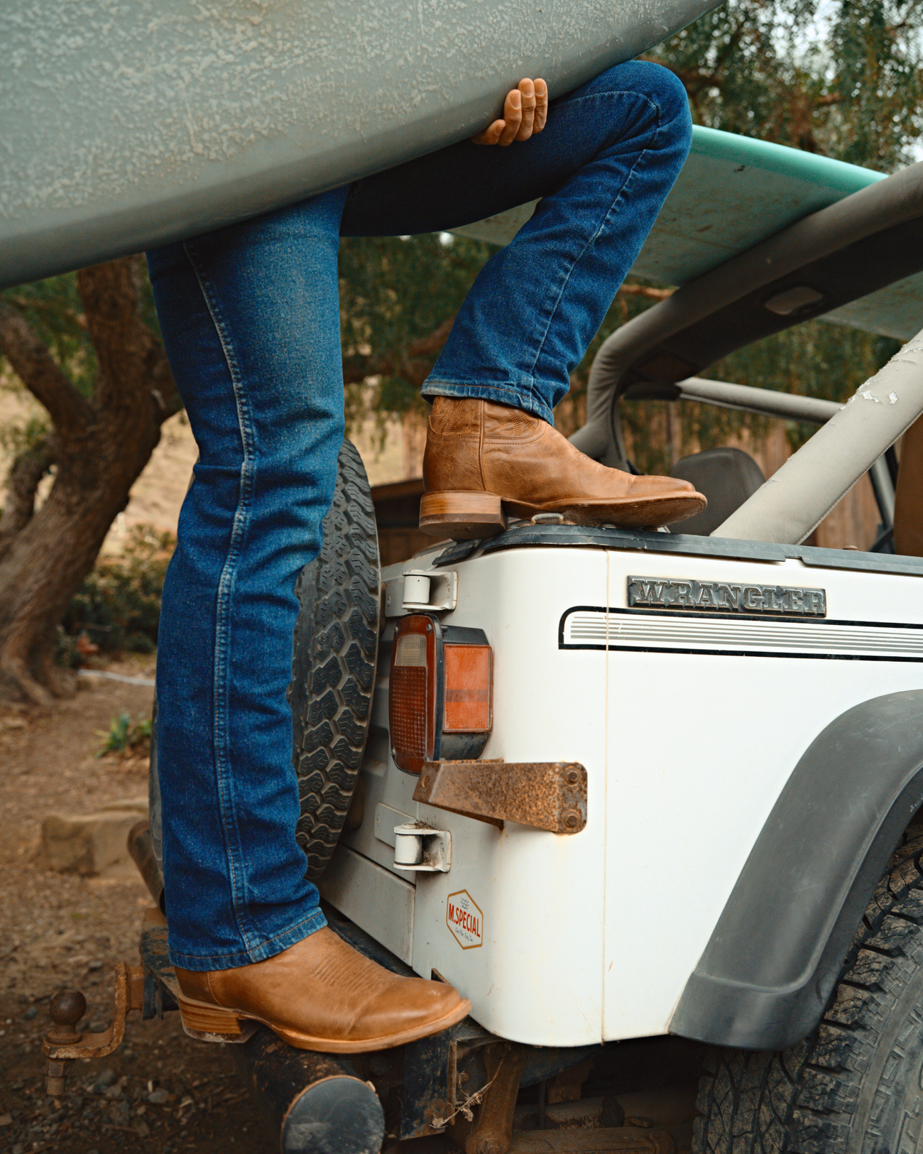 Person in jeans and cowboy boots climbing into a white Jeep, holding a surfboard.