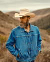 Man in denim jacket and cowboy hat stands in a mountainous landscape.