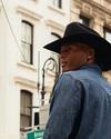 A person in a black cowboy hat and denim jacket stands on a city street with historic buildings in the background.