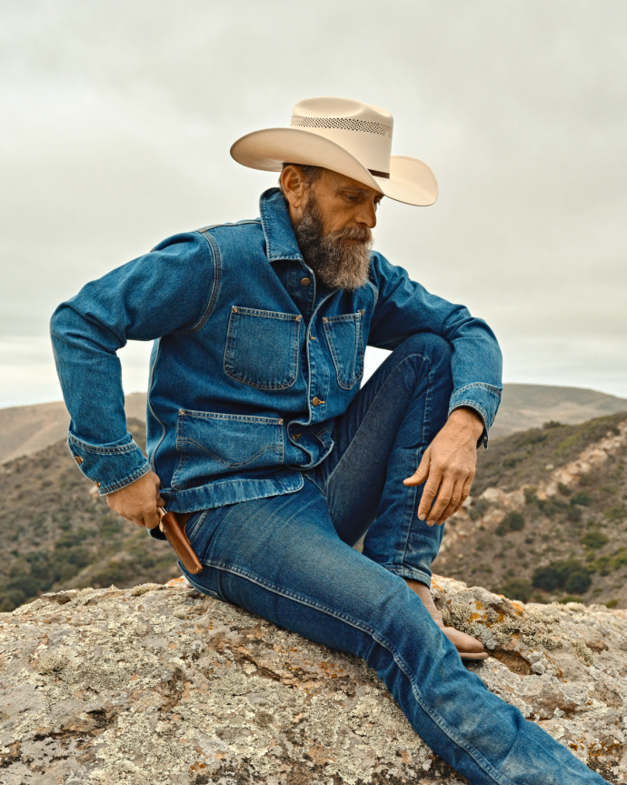 A person in a denim outfit and cowboy hat sits on a rock, holding a holstered item. Rugged landscape with hills in the background.