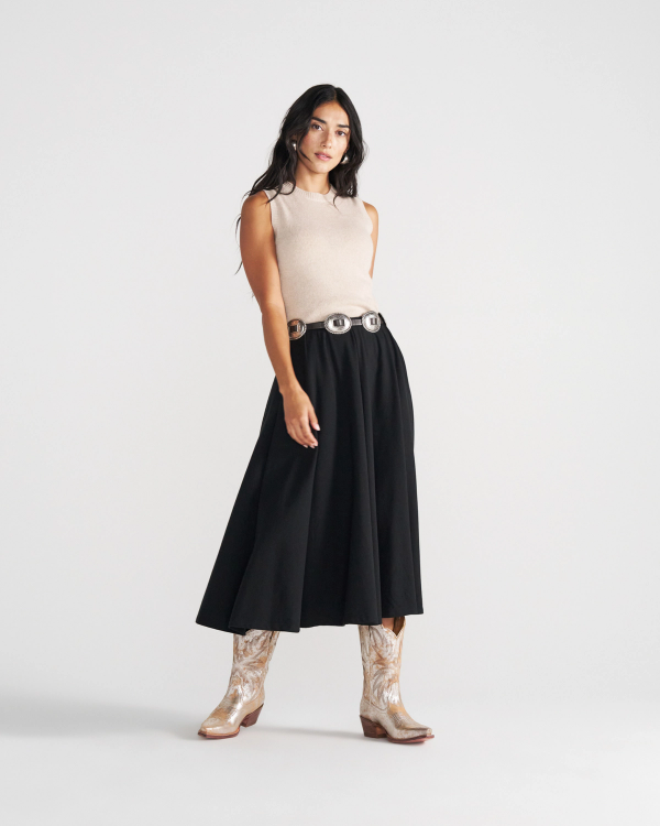 The Sadie in Platino Cowhide. A woman wearing a beige sleeveless top, a black midi skirt with a silver concho belt, and Platino Cowhide western-style boots.