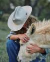A person in a cowboy hat and denim clothes hugs a light-colored donkey outdoors, holding the donkey’s head gently with both hands.