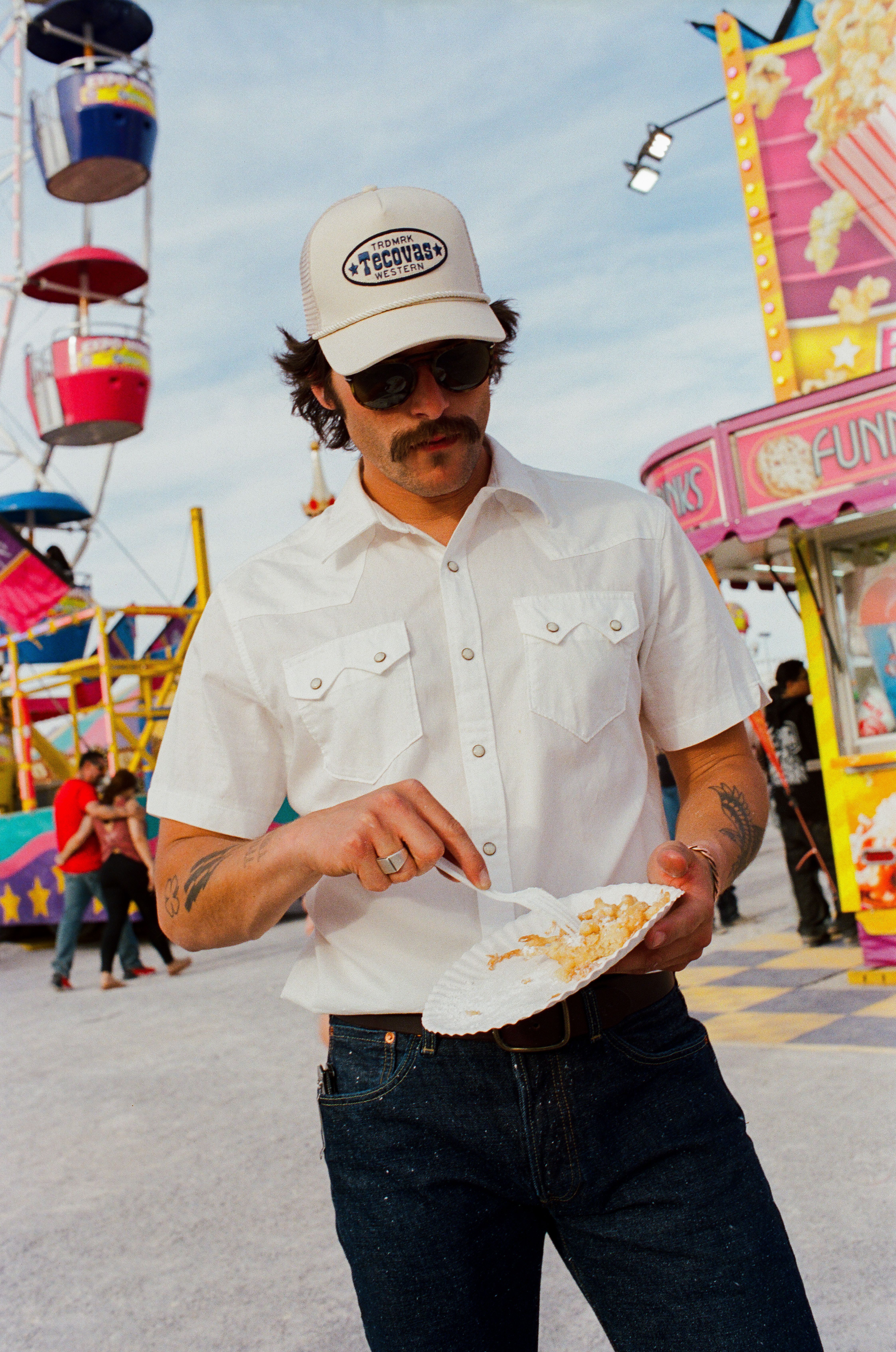 A man in sunglasses and a white shirt eats food from a paper plate at a carnival, with a Ferris wheel and food stands in the background.