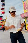 A man in sunglasses and a white shirt eats food from a paper plate at a carnival, with a Ferris wheel and food stands in the background.