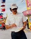 A man in sunglasses and a white shirt eats food from a paper plate at a carnival, with a Ferris wheel and food stands in the background.